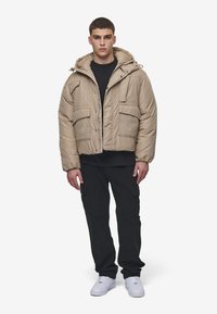 Beige dunjakke med hette, glidelås og fire frontlommer. Båret over en svart skjorte med mørke cargo-bukser og hvite joggesko.