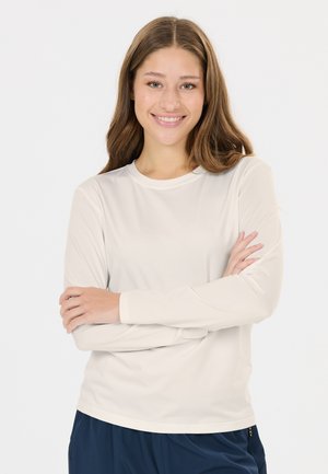 Jeune femme aux longs cheveux bruns sourit, portant une chemise blanche à manches longues et un pantalon bleu marine, les bras croisés.