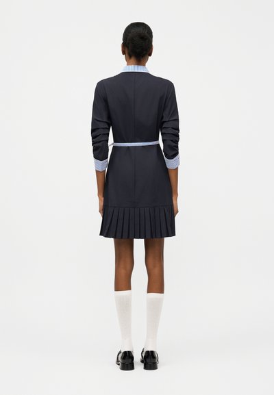 Ženska stoji obrnjena stran, oblečena v črno obleko z nagubanimi robovi, srajčne manšete z črtami, bele nogavice do kolen in črne čevlje na enobarvnem ozadju.