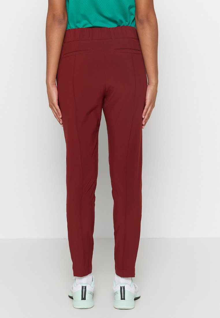 Pantalons bordeaux ajustés avec une taille plate, des poches arrières et une texture lisse. Portés avec des chaussures de sport claires.