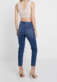 Bralette en dentelle beige clair avec un dos en V, associé à un jean en denim bleu taille haute et des chaussures à talons nus.