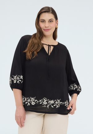 Kobieta ubrana w czarną bluzkę z białą kwiatową wyszywanką na rękawach i dole, zestawioną z beżowymi spodniami, uśmiechająca się do kamery.