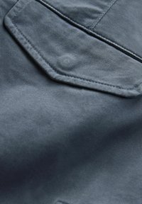 Teren de denim de culoare albastră închisă, cu o textură netedă. Prezintă un buzunar hexagonal distinct și detalii subtile de cusătură.