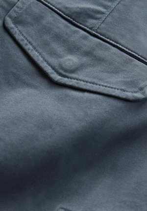 Denim w ciemnoniebieskim kolorze o gładkiej teksturze. Posiada charakterystyczną heksagonalną kieszeń i subtelne detale szycia.