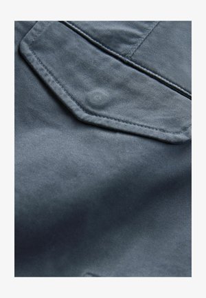 Tumši zila džinsu audums ar gludu tekstūru. Iezīmējas ar izteiktu sešstūru kabatu un subtiliem šuvumu elementiem.