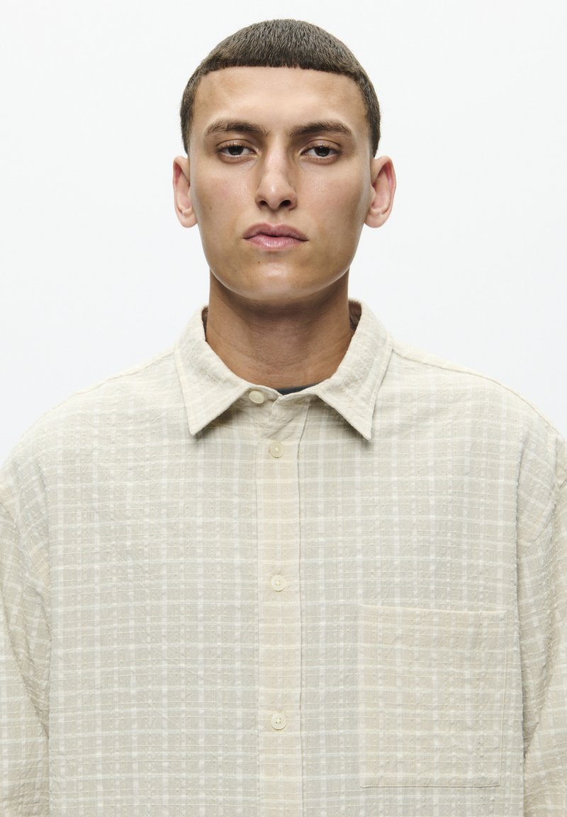Chemise à carreaux beige en tissu léger, avec une fermeture à boutons, un col pointu et une poche poitrine unique.