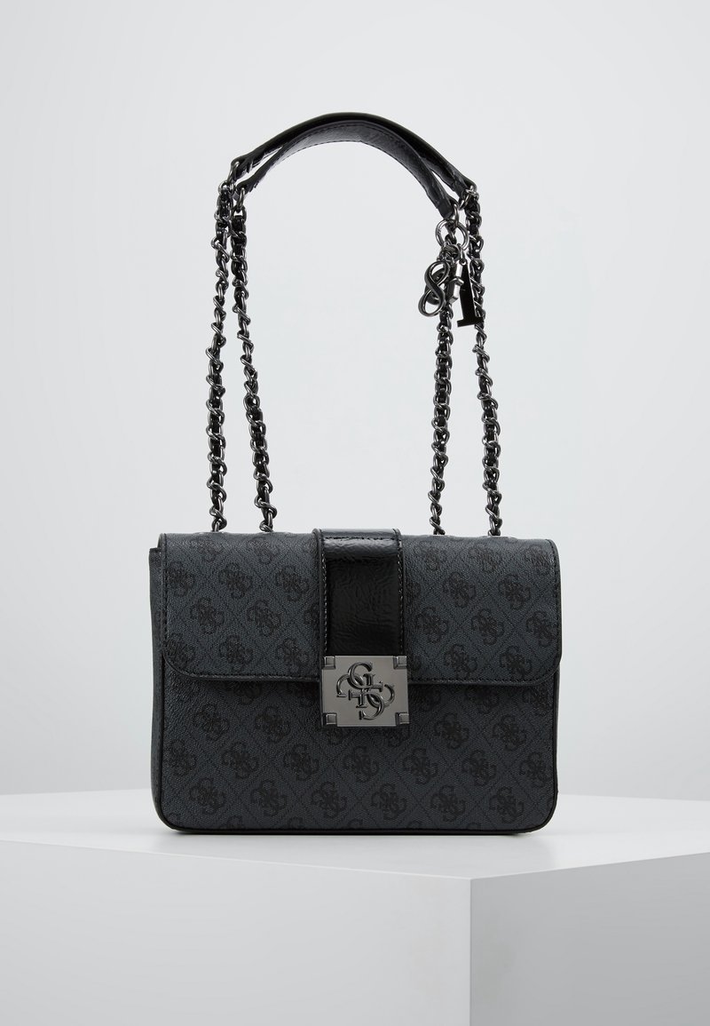 Sac à main de créateur noir avec bandoulière chaîne, surface à motifs et fermoir logo métallique à l'avant sur un socle blanc, fond uni.