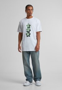 White cotton t-shirt featuring three overlapping green stars printed vertically. Paired with loose-fitting faded blue jeans and white sneakers.