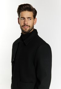 Schwarzer Wollmantel mit Kragen, einer einzelnen Tasche und strukturiertem Finish. Modell trägt einen schwarzen Rollkragenpullover. Minimalistisches Design mit subtiler Markenkennzeichnung.