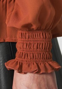 Rostfärgad blus med genomskinligt, strukturerat tyg och smockade ärmslut med volanger i kanten. Nederdelen är figursydd med en rak fåll.