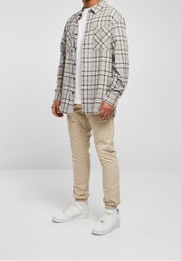 Uomo che indossa pantaloni cargo beige, sneakers bianche, t-shirt bianca e camicia a quadri grigia con bottoni, in piedi su uno sfondo grigio.