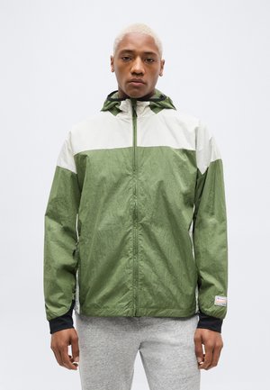 Άτομο με κοντά ξανθά μαλλιά, που φοράει πράσινο και λευκό φούτερ με κουκούλα και φερμουάρ και ανοιχτό γκρι παντελόνι, όρθιο μπροστά σε απλό φόντο.