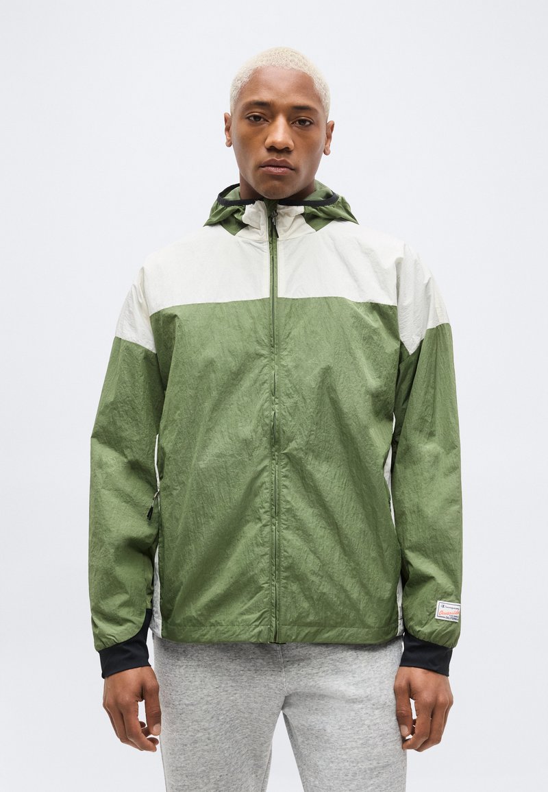 Lühikeste blondide juustega inimene, kes kannab rohevalget kapuutsiga lukuga jope ja helehallid pükse, seisab ühevärvilise tausta ees.