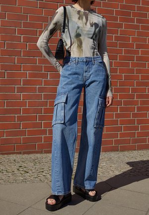 Jeans cargo anchos azules con grandes bolsillos laterales; combinados con una blusa de manga larga ajustada con estampado y sandalias de plataforma negras.