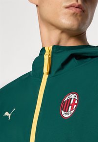 Grøn jakke med gul lynlås, prydet med et hvidt Puma-logo og et rundt, rødt AC Milan-emblem med sorte striber og numeriske detaljer.