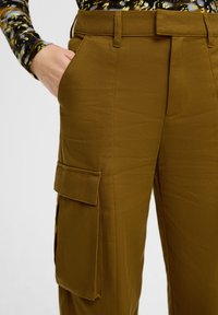 Bruine, gestructureerde cargo broek met een vlakke voorkant, zijzakken en een grote zak op de dij. Ontworpen voor functionaliteit en comfort.