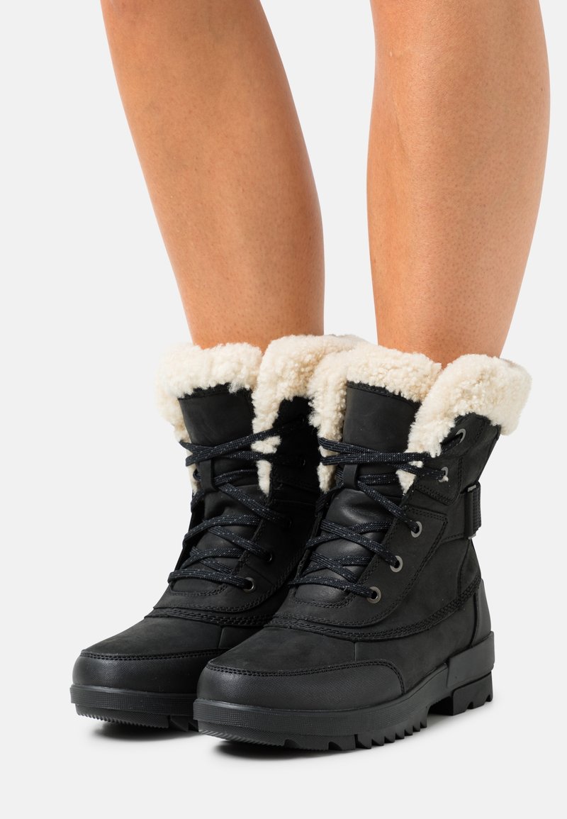 Bottes d'hiver noires avec semelles en caoutchouc épaisses et doublure en polaire crème portées sur des jambes nues.