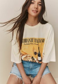 Crème oversized t-shirt met een vintage Italiaanse grafiek, met een gele achtergrond en voedsel/wijn motieven. Gecombineerd met versleten denim shorts.