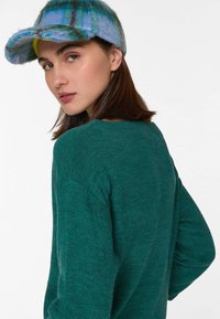 Grüner Strickpullover mit strukturiertem Finish. Das Modell trägt eine blau-grüne Karo-Mütze mit einem geschwungenen Schirm und hellen Akzenten.