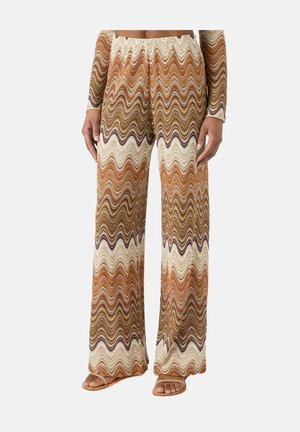 Pantalon à jambes larges avec un motif zigzag ondulé dans des tons marron, beige et blanc, porté avec des sandales ouvertes couleur camel.