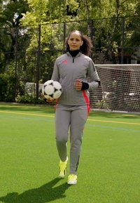 Chándal deportivo gris con acentos rojos, sosteniendo un balón de fútbol en blanco y negro, sobre césped verde, con una portería de fútbol de fondo.