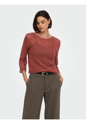 Femme aux cheveux courts et foncés portant des boucles d'oreilles en anneaux dorés, un haut en dentelle tricotée couleur rouille et un pantalon marron à jambes larges, les mains dans les poches.
