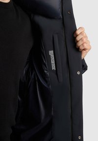 Chaqueta acolchada de color azul oscuro con un interior de nailon suave, que cuenta con cierre de cremallera, botones a presión y un bolsillo interior oculto que muestra las instrucciones de cuidado.