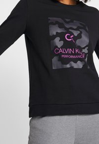 Musta huppari, jossa on kamouflagekuvio ja vaaleanpunaista tekstiä, jossa lukee "Calvin Klein Performance". Rentomalli. Sileä pinta.
