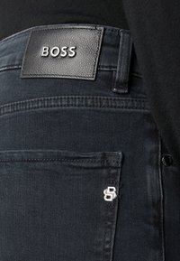 Slim-fit tume denim teksad, millel on tekstuuriga musta nahkide etikett, kus on sõna "BOSS". Hõbedased aksessuaari detailid on nähtavad tasku lähedal.