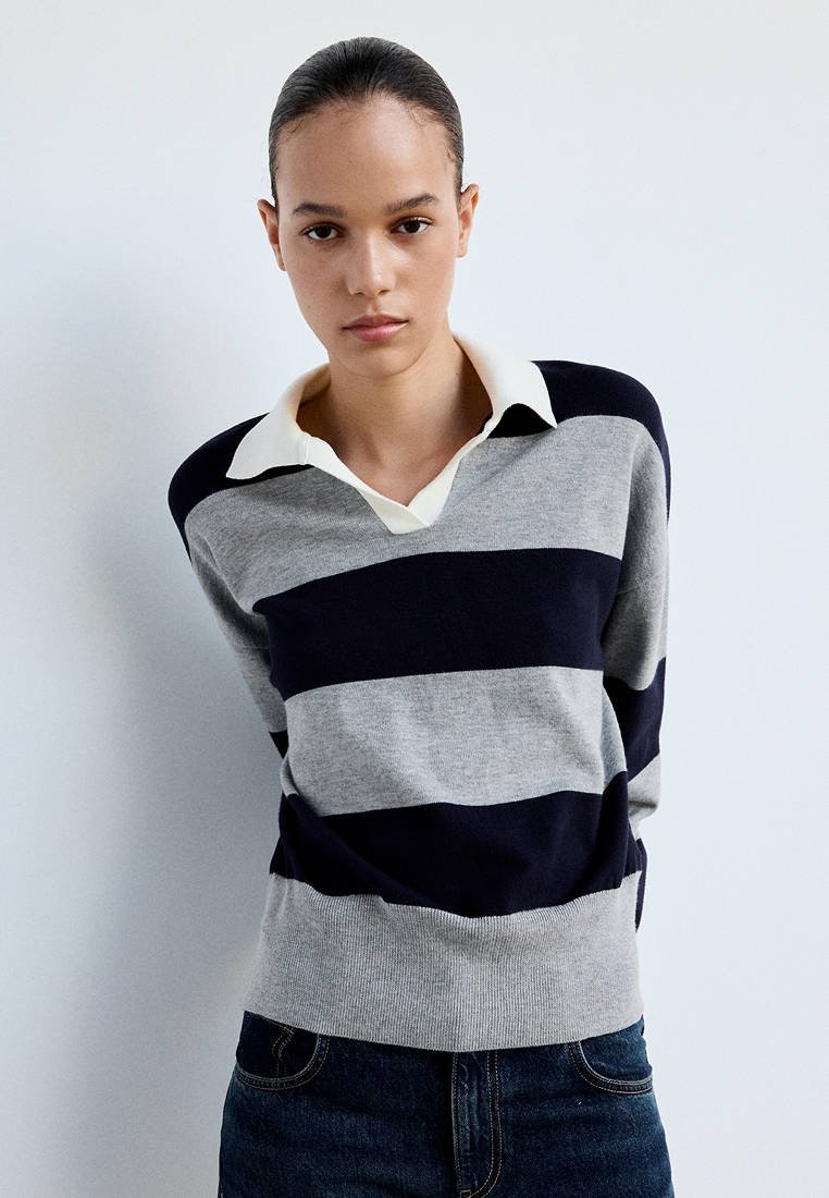 Jeune personne aux cheveux attachés, portant un pull rayé gris et bleu marine sur une chemise blanche à col et un jean bleu foncé.