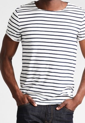 Mężczyzna w białej koszuli z krótkim rękawem z czarnymi poziomymi paskami i ciemnoniebieskich dżinsach, z rękami opartymi w przednich kieszeniach.