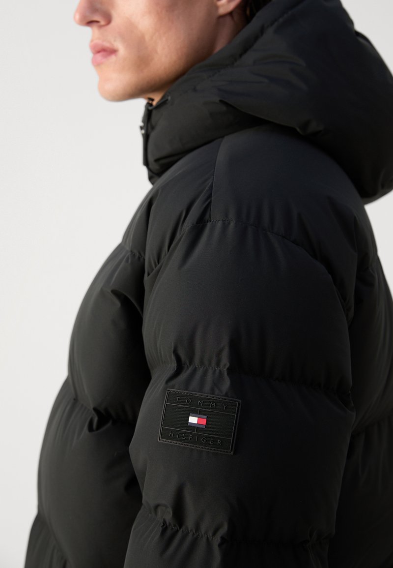 Chaqueta puffer negra con una capucha prominente, que presenta una superficie texturizada, segmentos acanalados y un parche de logo rectangular en la manga.