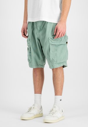 Uomo che indossa pantaloncini cargo verde chiaro, calzini bianchi e scarpe da ginnastica bianche, in piedi contro uno sfondo semplice.