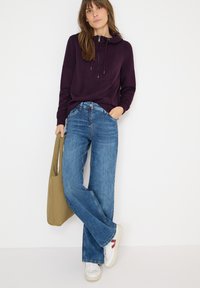 Felpa viola con tasca frontale e cordino; jeans a gamba larga azzurri; sneakers bianche con dettagli rossi; borsa a tracolla verde oliva.