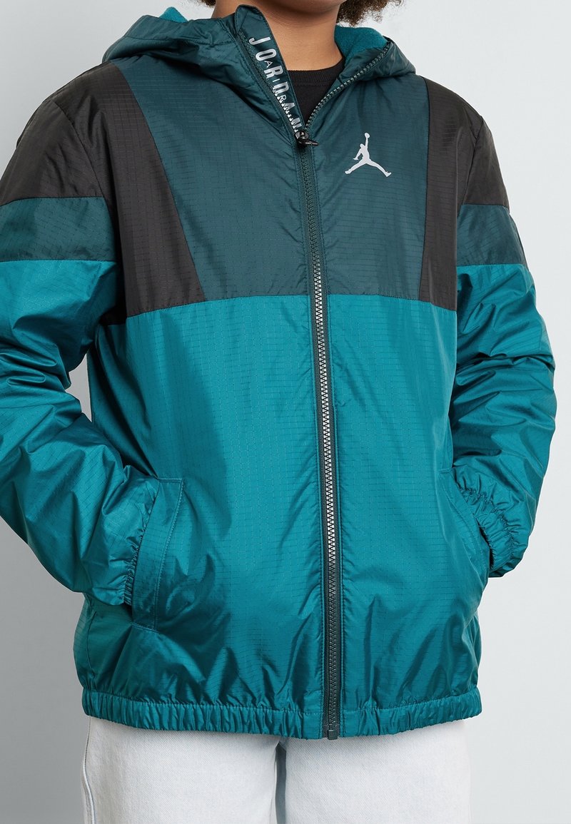 Teal og sort zip-up jakke med hætte, med en struktureret overflade, elastiske manchetter og et hvidt logo på brystet.