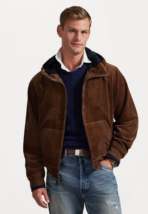 Giacca in corda marrone con cappuccio, caratterizzata da una chiusura lampo frontale, due grandi tasche e una fodera a quadri neri. Indossata sopra un maglione blu navy.
