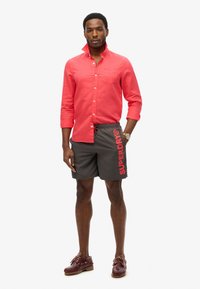 Rode lange mouwen shirt, grijze zwembroek met "SUPERDRY" logo, bruine bootschoenen. Model staat rechtop tegen een witte achtergrond.