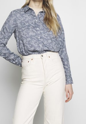 Mujer con una camisa azul de flores abotonada, metida en unos vaqueros blancos de tiro alto, posando con una mano en la cadera sobre un fondo liso.