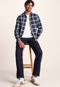 Plaid flannel shirt in navy, teal, and white, paired with dark denim jeans and white sneakers with black stripes. Seated on a wooden stool.