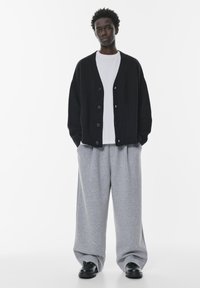 Cardigan negru tricotat cu margini zimțate, nasturi mari și o croială lejeră, purtat deasupra unui tricou alb și a unor pantaloni de trening largi gri. Pantofi negri.