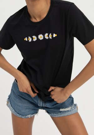 Person wearing black T-shirt with seven white daisies in a row on the chest and blue denim shorts with frayed edges.
