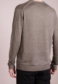 Homme portant un pull en maille taupe à manches longues avec poignets et ourlet côtelés, debout de dos face à la caméra sur un fond uni.