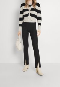 Cárdigan de rayas en blanco y negro con botones, combinado con pantalones negros de dobladillo partido y un bolso crema. Botines de tacón beige completan el conjunto.