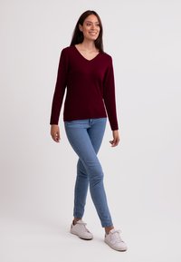 Jersey burdeos de cuello en V con mangas largas, combinado con jeans ajustados azul claro y zapatillas blancas. Textura suave, diseño ajustado.