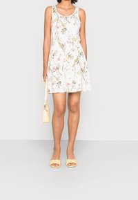 Floral-patterned, sleeveless dress in white with pink and yellow flowers. Paired with yellow heeled sandals and a small handbag.