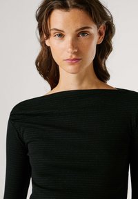 Μαύρο τοπ με μακριά μανίκια και ριμπ ύφανση, με φαρδύ ντεκολτέ τύπου βάρκα και πιέτες στους ώμους. Το ύφασμα έχει απαλή, υφή με υφή.