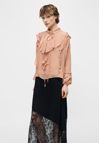 Γυναίκα με ανοιχτό ροζ μπλούζα με βολάν και μακριά μανίκια και μαύρη ασύμμετρη φούστα με δαντελένιο επάνω στρώμα, που στέκεται μπροστά σε απλό φόντο.