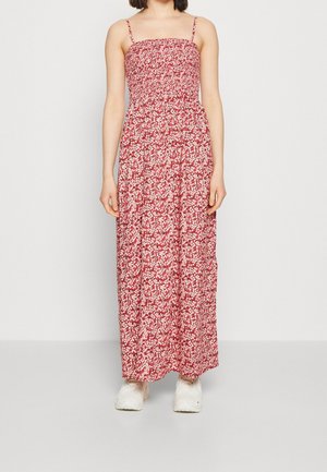 Kobieta w długiej czerwonej sukience w kwiaty na cienkich ramiączkach i białych trampkach, stojąca na tle jednolitego jasnoszarego tła.