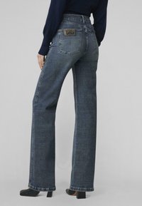 Granatowe dżinsy o wysokim stanie, w kroju szerokich nogawek, z naszywaną kieszenią z tyłu z logo. Teksturowana tkanina, minimalne detale.