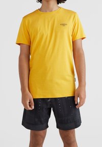 Camiseta amarilla de cuello redondo y mangas cortas; presenta un pequeño logo negro. Combinada con pantalones cortos negros con un diseño estampado.